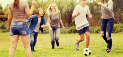 Group of carefree teenagers having fun and kicking football in park on summer day