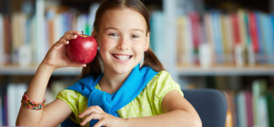 Portrait of happy schoolgirl with big red apple looking at camera in library